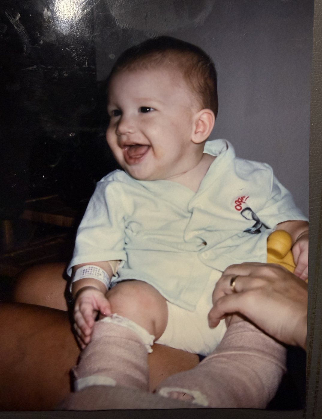 Baby Heath laughing on a lap, with casts on both legs and a hospital band on his wrist