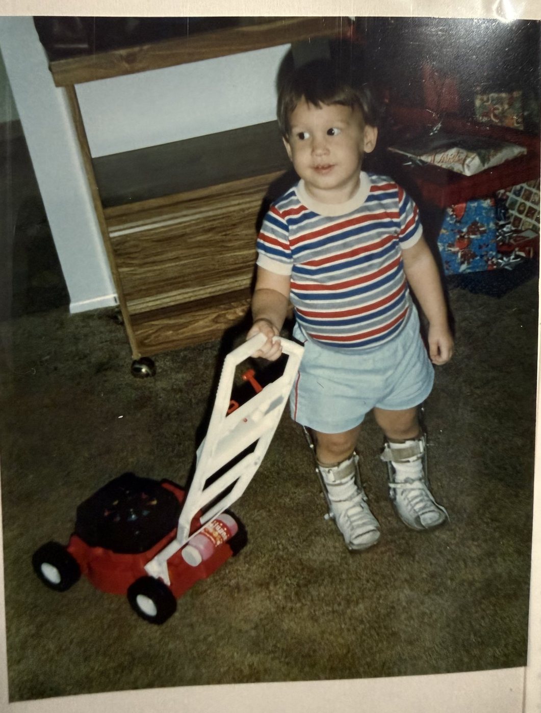 Toddler standing on carpet pushing a toy lawnmower, wearing white shoes with metal uprights up both legs