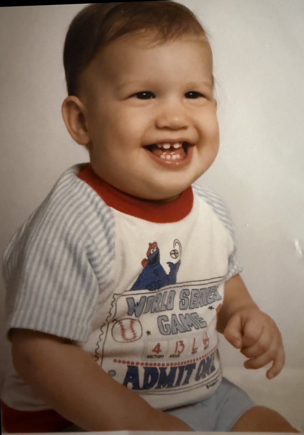 Grinning toddler in a studio portrait wearing a baseball ticket T-shirt