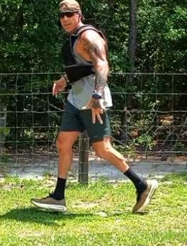 Heath mid-stride on grass in a vest, cap and sunglasses, both feet in frame