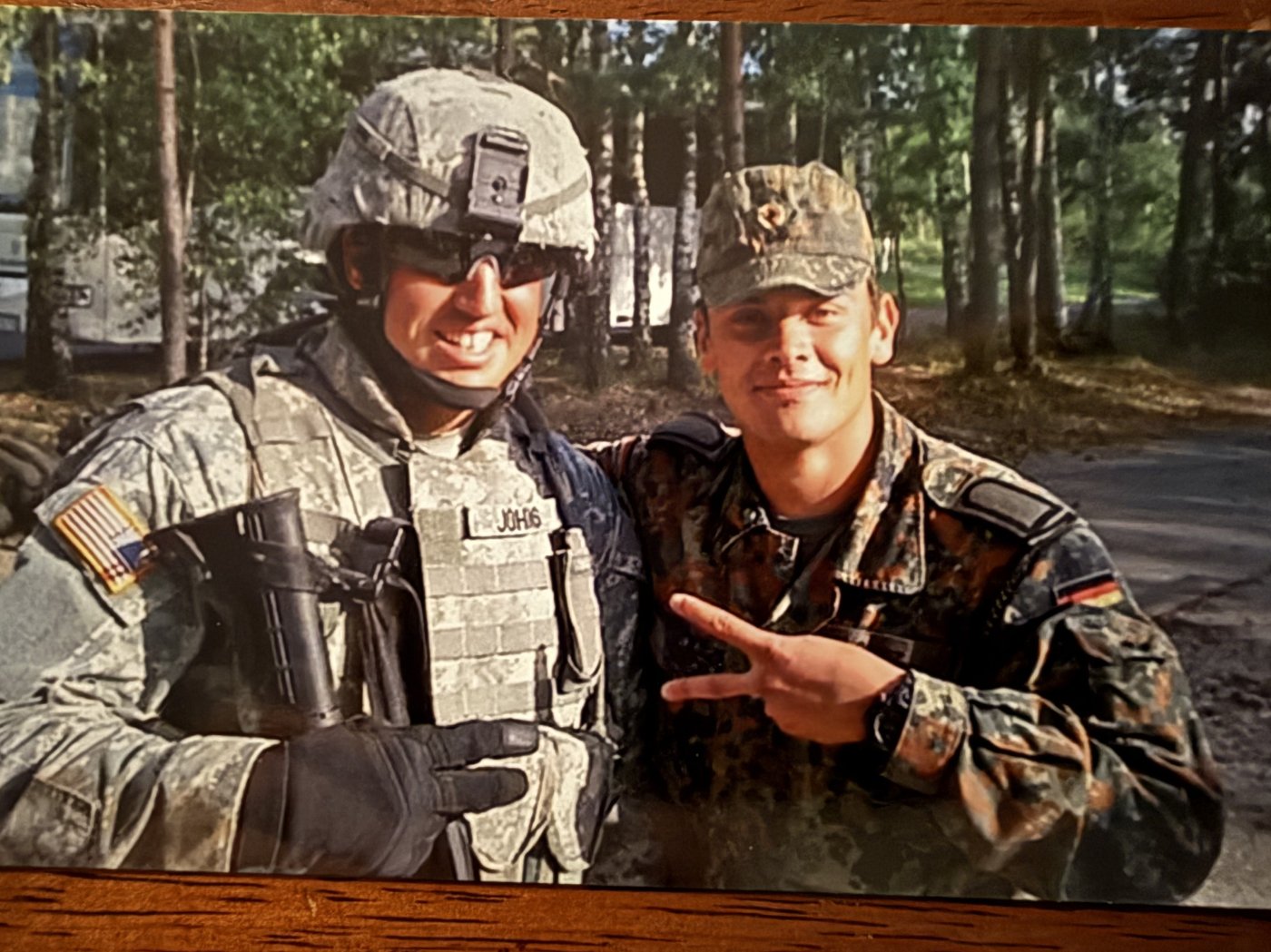 Soldier in US combat uniform and helmet standing beside a soldier in German camouflage on a wooded road